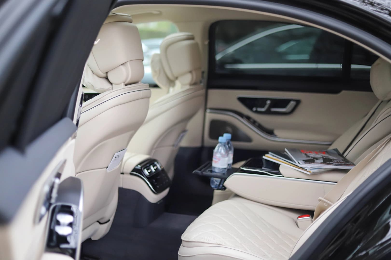Interior of a car showcasing cream-colored, luxurious leather seats with detailed stitching. A bottle of water and magazines are placed on one of the seats. The interior design includes sleek black and cream contrasts, with modern controls visible on the rear armrest. Ideal for luxury car rental services.
